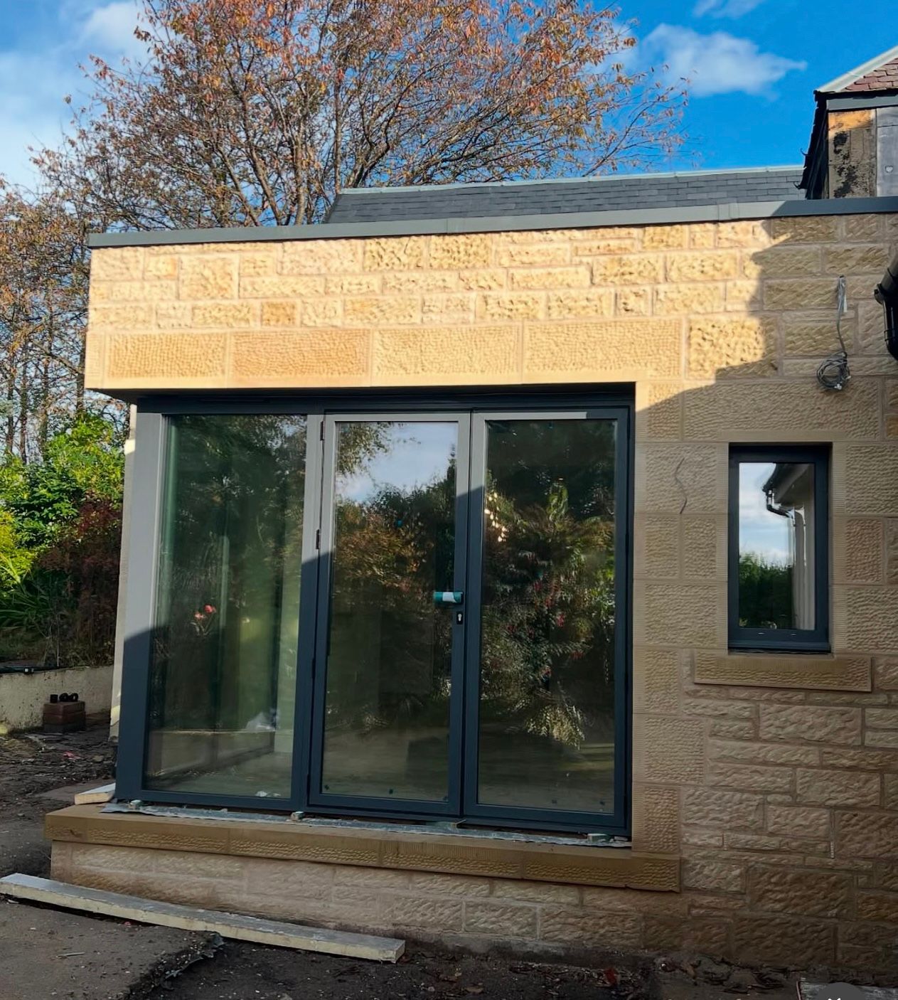 Bright sandstone stonework on modern extension with large grey framed glass door and small window.